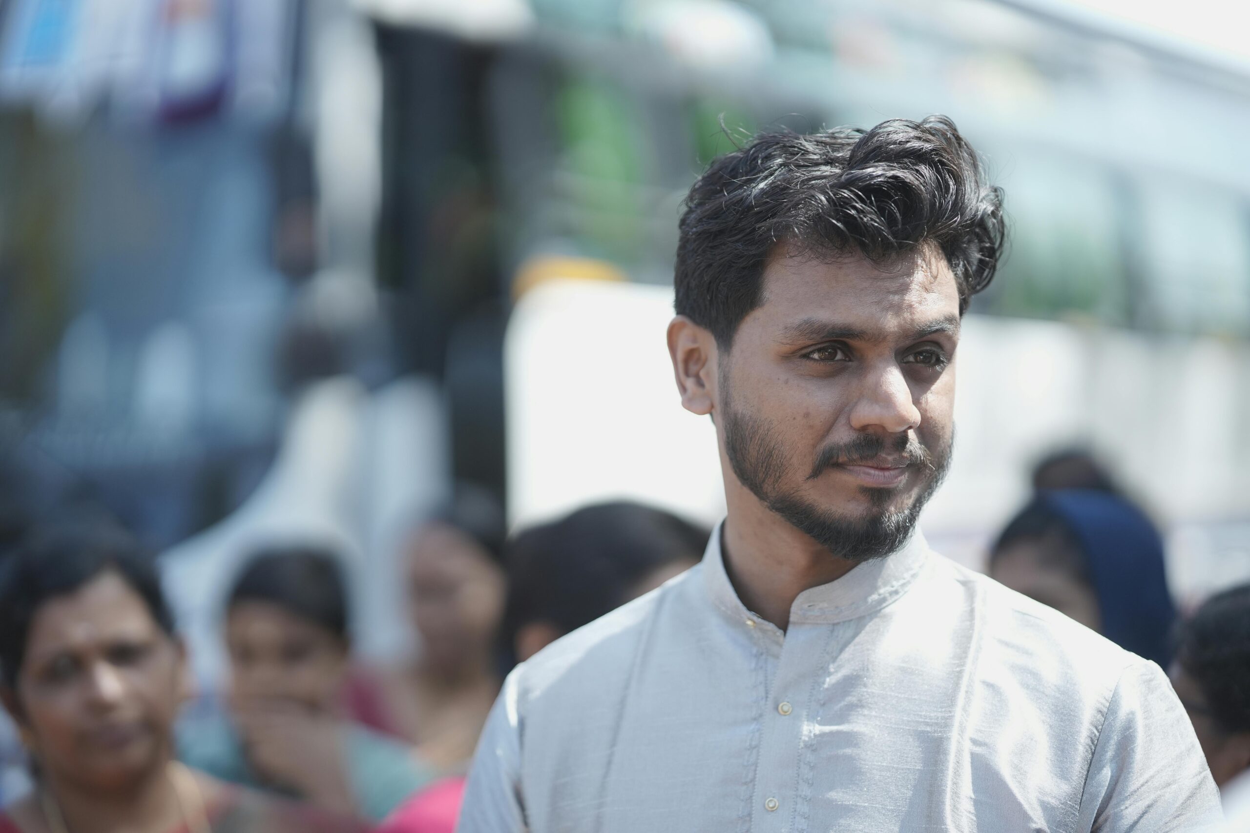 A thoughtful man in traditional attire during an outdoor event, surrounded by people.